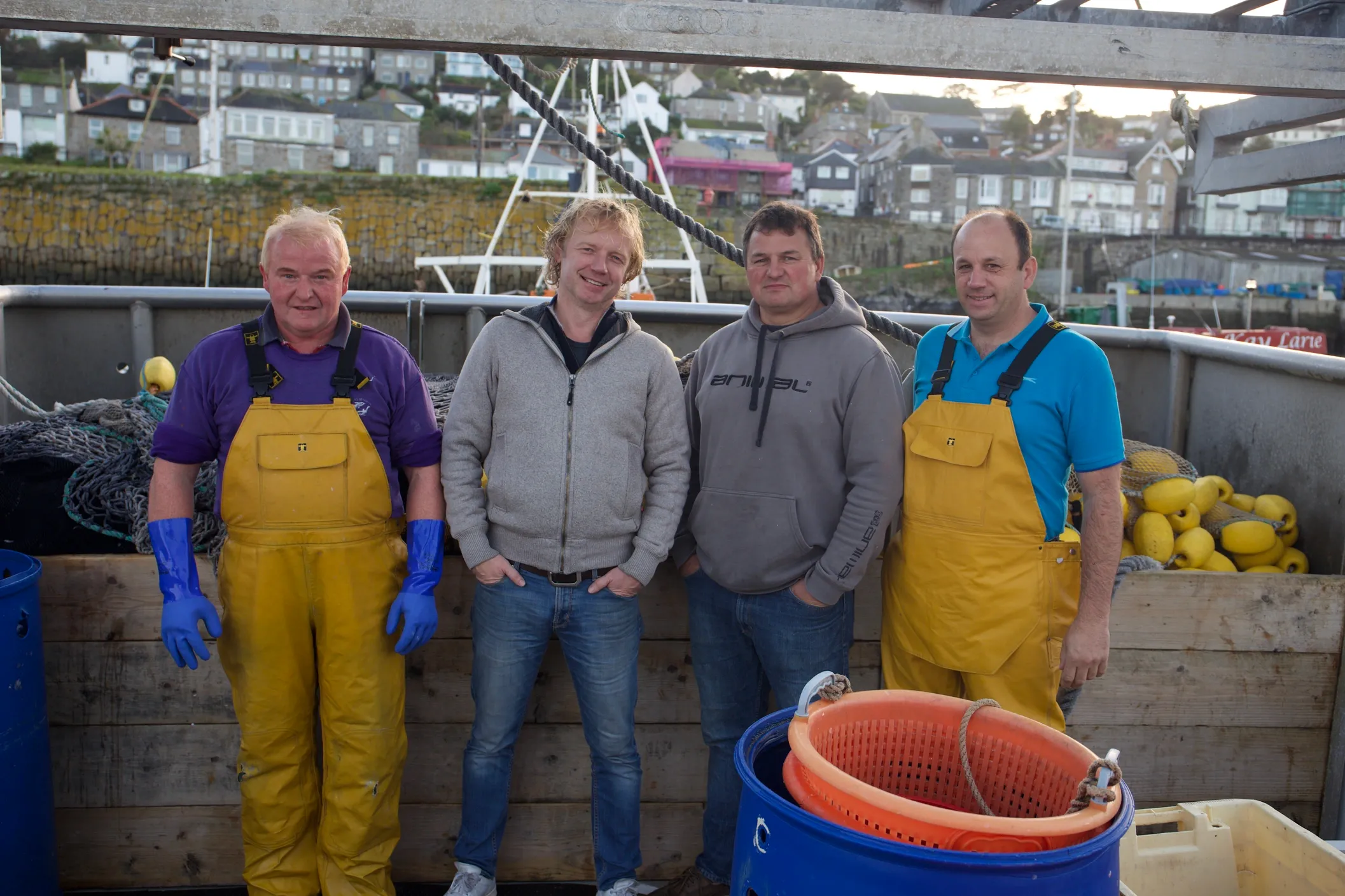 Four fishermen in waders and casual clothes stand on a boat with nets and buoys.