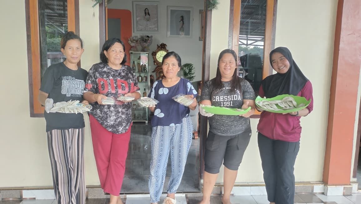 Five smiling women holding plates of freshly prepared food in front of a house.