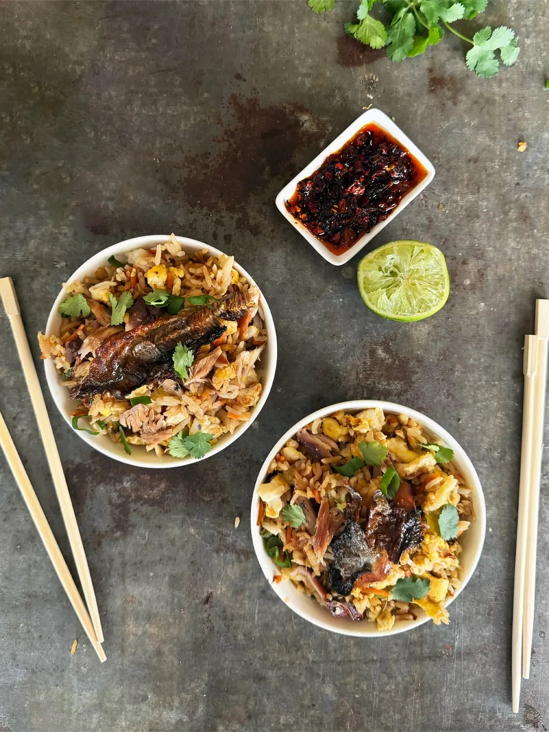 Two bowls of fried rice with meat, herbs, chopsticks, sauce, and a lime wedge.