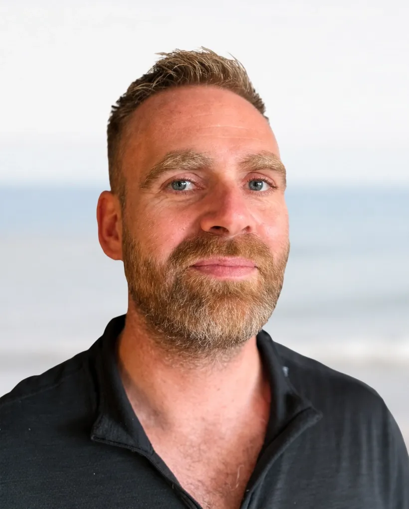 A bearded man with short hair smiles slightly in front of a blurred ocean background.