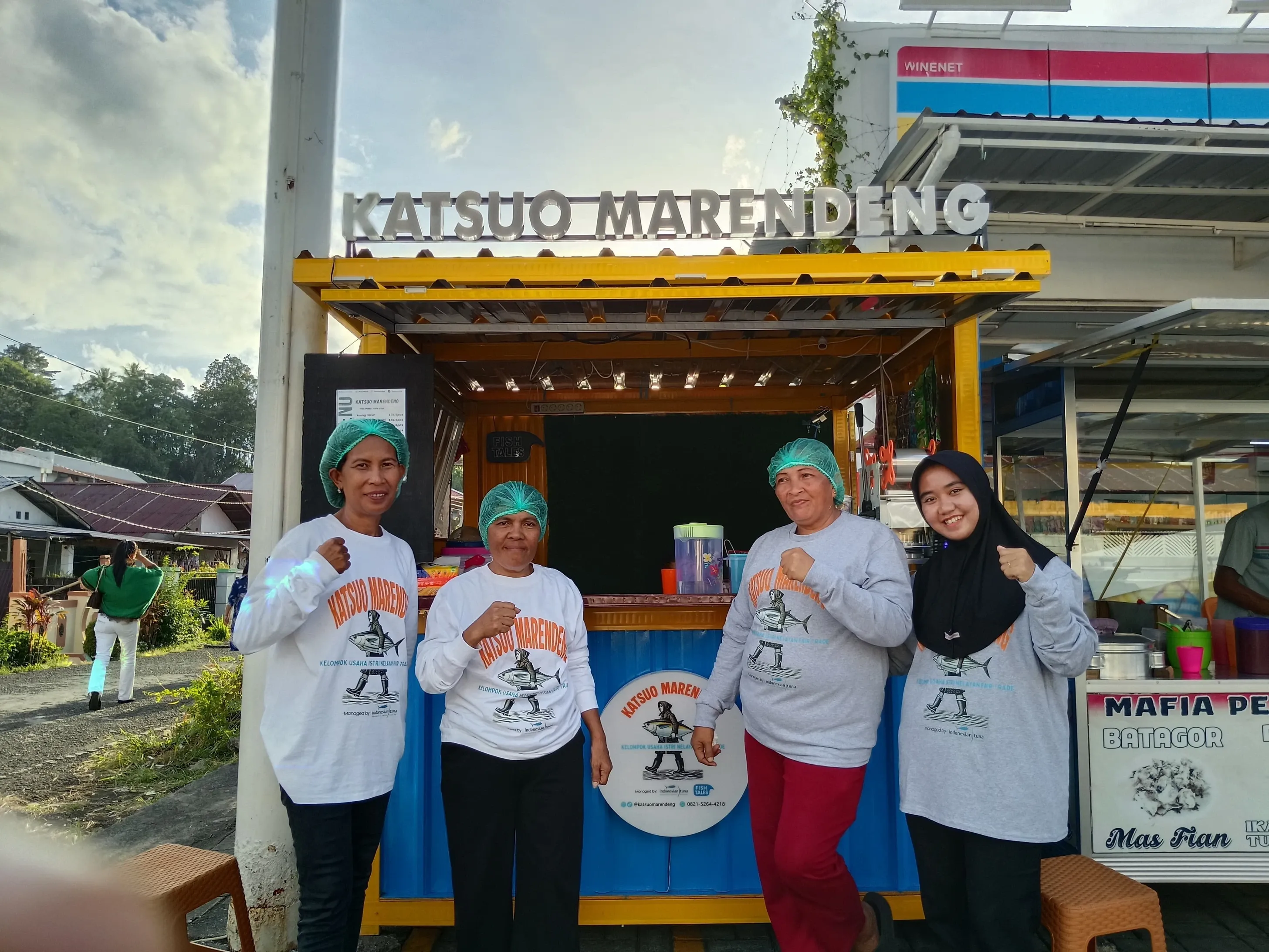 Four women in matching uniforms and hairnets pose proudly in front of a 'Katsuo Marendeing' food stall.