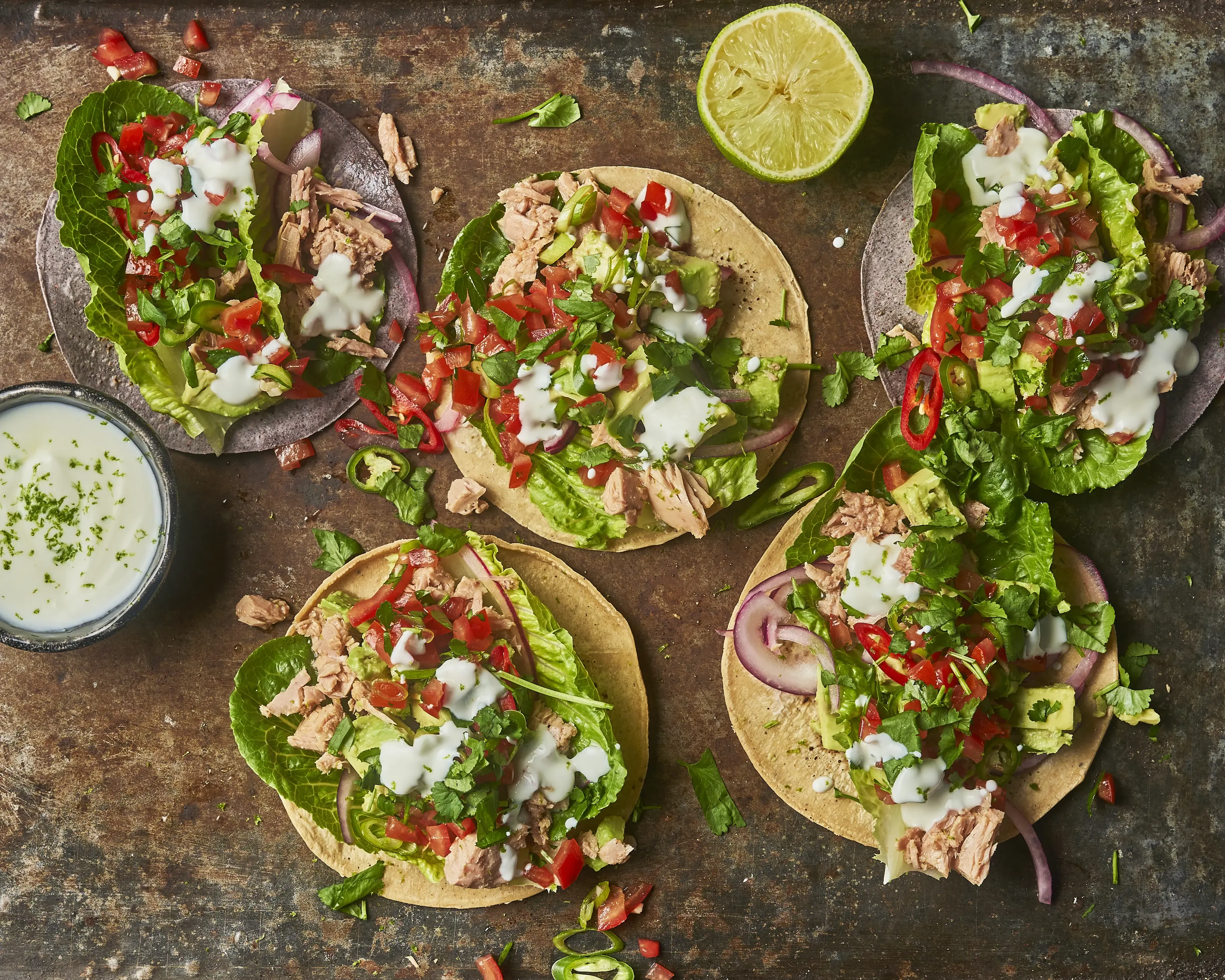 Fresh tacos with fish, avocado, lettuce, tomato, and lime on a rustic surface.
