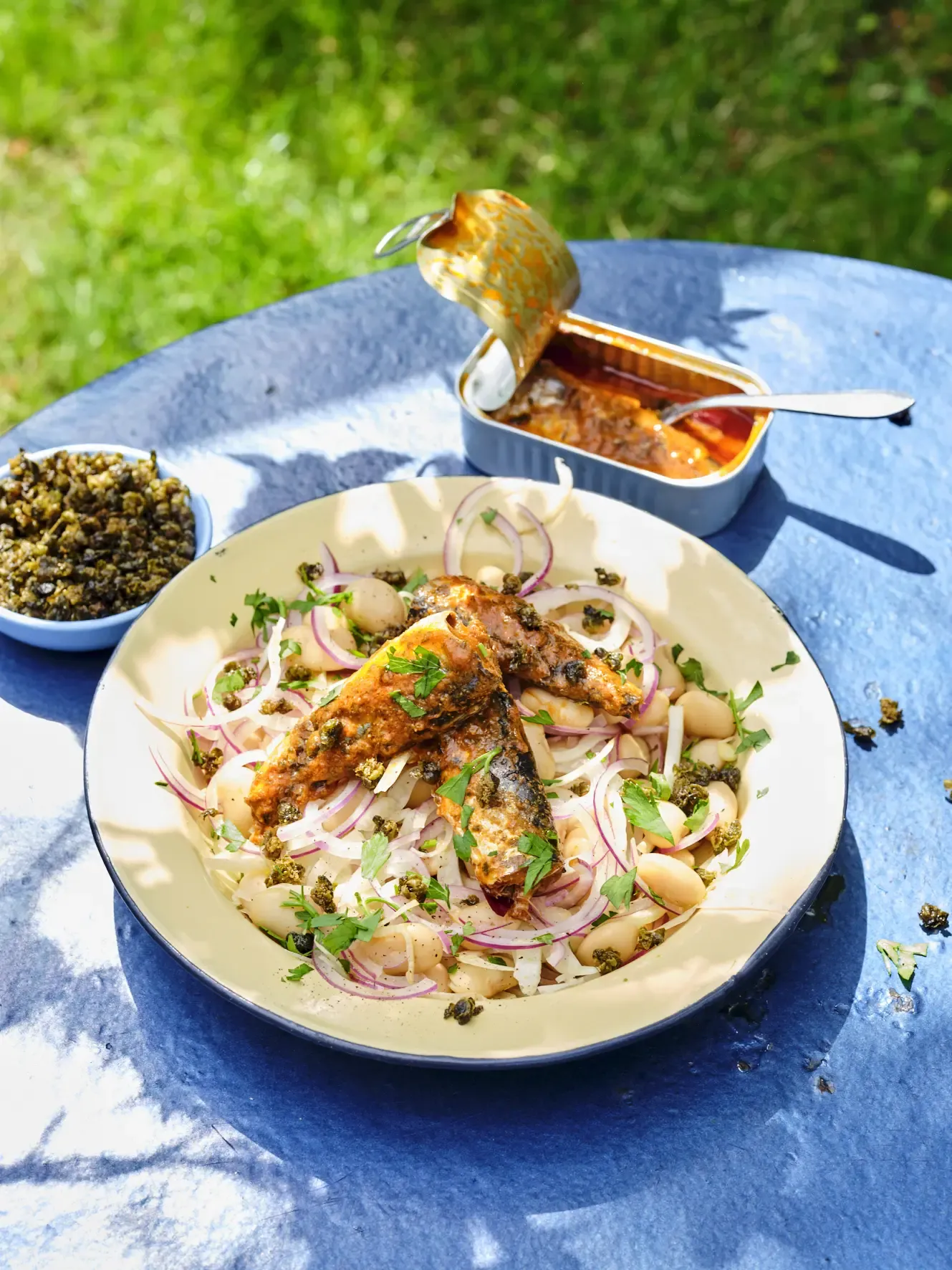 A sardine bean salad with red onions and capers is served on a blue table outdoors.